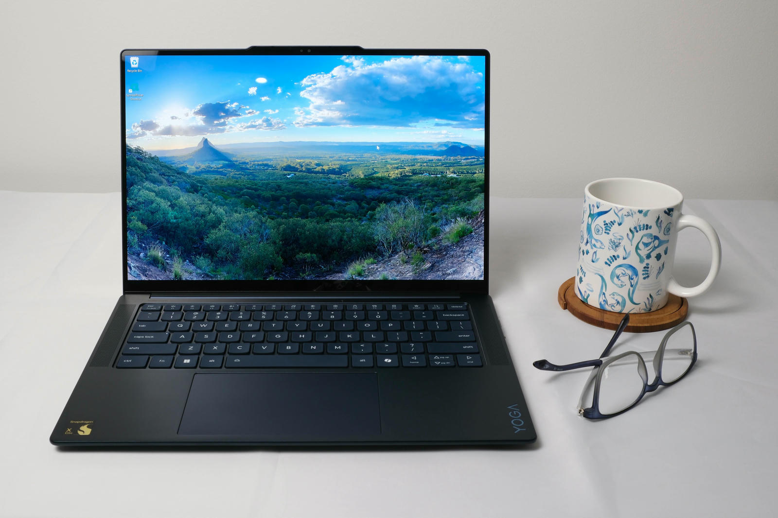 Photo of the Yoga 7x laptop open on a desk showing the Glass House Mountains on the desktop. To the right of the laptop is a coffee mug and a pair of glasses.